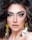 A close-up portrait of a person with striking blue eyes, full, arched eyebrows, and long, voluminous eyelashes. The makeup is sophisticated, highlighting the eyes and lips, with a soft nude lipstick. The hair is styled in loose, wavy curls. Large, shiny, circular earrings with gold detailing add a glamorous touch.