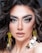 A close-up portrait of a person with striking blue eyes, full, arched eyebrows, and long, voluminous eyelashes. The makeup is sophisticated, highlighting the eyes and lips, with a soft nude lipstick. The hair is styled in loose, wavy curls. Large, shiny, circular earrings with gold detailing add a glamorous touch.