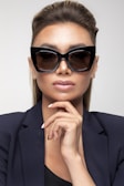 A person wearing large, stylish black sunglasses and a dark blazer with a confident expression. The person is touching their chin with manicured fingers featuring white nail polish, and their hair is elegantly styled.