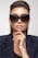 A person wearing large, stylish black sunglasses and a dark blazer with a confident expression. The person is touching their chin with manicured fingers featuring white nail polish, and their hair is elegantly styled.