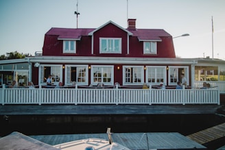 A red wooden house with white trim features a large terrace where several people are seated, possibly enjoying a meal or drink. The building has a classic Scandinavian architectural style with multiple windows and a sloped roof. A white picket fence encloses the terrace, and the setting suggests a quaint and inviting atmosphere.