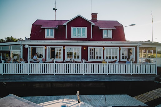 A red wooden house with white trim features a large terrace where several people are seated, possibly enjoying a meal or drink. The building has a classic Scandinavian architectural style with multiple windows and a sloped roof. A white picket fence encloses the terrace, and the setting suggests a quaint and inviting atmosphere.