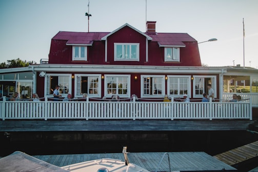 A red wooden house with white trim features a large terrace where several people are seated, possibly enjoying a meal or drink. The building has a classic Scandinavian architectural style with multiple windows and a sloped roof. A white picket fence encloses the terrace, and the setting suggests a quaint and inviting atmosphere.