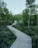 A winding wooden pathway leads through lush, green vegetation, surrounded by tall trees. A person wearing a red jacket and carrying a backpack walks along the path, adding a sense of exploration and adventure. The scene conveys a tranquil and serene natural environment.
