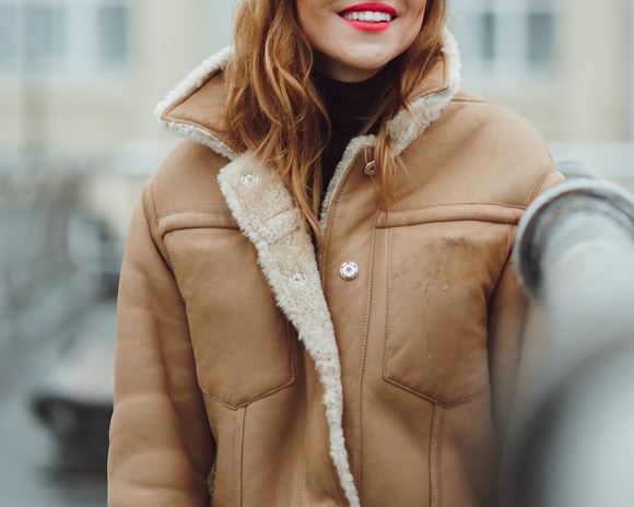 A person wearing a tan shearling jacket with a soft, fuzzy collar and visible buttons. The focus is on the upper body, with a backdrop that appears to be outdoors. The individual has long, wavy hair and is smiling with bright red lipstick.