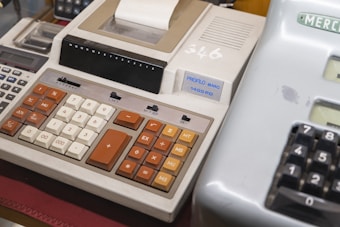 A vintage adding machine with large, tactile buttons in shades of orange, brown, and white. It has a paper roll for printing calculations and several switches and function keys.
