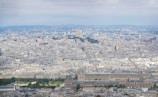 A panoramic cityscape showing a dense urban area with a mix of historical and modern architecture. A notable basilica sits atop a hill in the distance, surrounded by classical buildings. The landscape is dotted with lush green parks, and a large Ferris wheel is visible prominently in the central area.