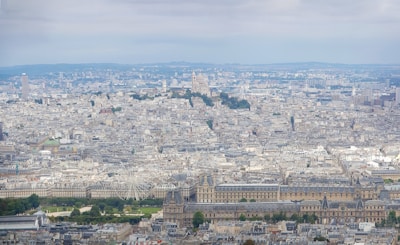 A panoramic cityscape showing a dense urban area with a mix of historical and modern architecture. A notable basilica sits atop a hill in the distance, surrounded by classical buildings. The landscape is dotted with lush green parks, and a large Ferris wheel is visible prominently in the central area.