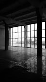 A large, empty industrial space with concrete floors and high ceilings. The right side features expansive windows, through which an industrial landscape, including cranes and ships, can be seen. The interior is dimly lit, creating reflections on the polished floor.