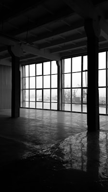 A large, empty industrial space with concrete floors and high ceilings. The right side features expansive windows, through which an industrial landscape, including cranes and ships, can be seen. The interior is dimly lit, creating reflections on the polished floor.