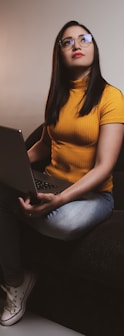 woman in yellow shirt and blue denim jeans sitting on black sofa using macbook