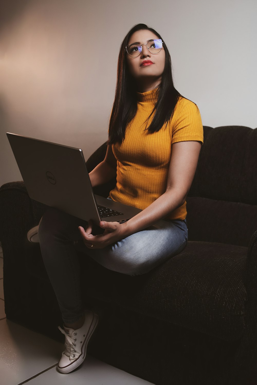 Frau in gelbem Hemd und blauer Jeans sitzt mit dem MacBook auf schwarzem Sofa