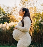 A pregnant woman standing outdoors in a lush, green environment with trees and grasses. She is wearing a textured cream-colored sweater and looking upwards with a serene expression. The sunlight filters through the trees, creating a warm, golden glow.