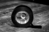 A black and white photograph shows a large roll of tape lying on its side on a wooden surface, with a smaller rolled-up object perfectly aligned inside it. The background is blurred, drawing attention to the rolls of tape.