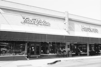 A vintage storefront of a hardware store with an awning and signage that reads 'True Value Hardware'. The building features large windows displaying various goods, with a classic design.
