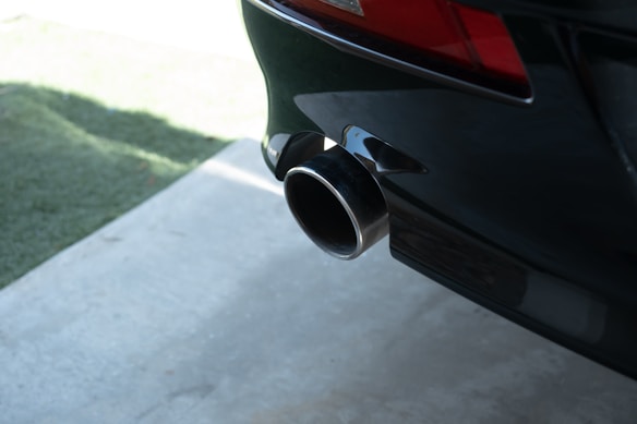 A close-up view of a car's exhaust pipe extending from the rear of the vehicle. The exhaust is clearly polished and reflects light. The car is parked on a concrete surface, with some grass visible in the background.