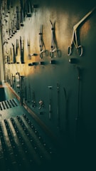 A wall-mounted display of various metal tools, possibly surgical or repair instruments, arranged in rows with hooks. The lighting casts long shadows, giving the scene a vintage and slightly eerie atmosphere.