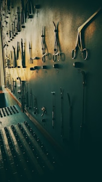 A wall-mounted display of various metal tools, possibly surgical or repair instruments, arranged in rows with hooks. The lighting casts long shadows, giving the scene a vintage and slightly eerie atmosphere.