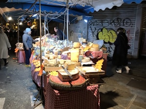 A street market stall is filled with a variety of cheeses displayed on red and white checkered tablecloths. Two people interact at the stall under a blue canopy during nighttime. The surrounding walls are covered in graffiti, adding an urban atmosphere to the scene.