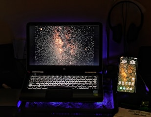 A laptop and a smartphone are displayed side by side on a dark surface. Both screens show an image of a starry sky, possibly the Milky Way. The laptop's keyboard is illuminated with a white backlight, adding a soft glow to the scene. A pair of headphones is visible in the background, hanging on the wall.