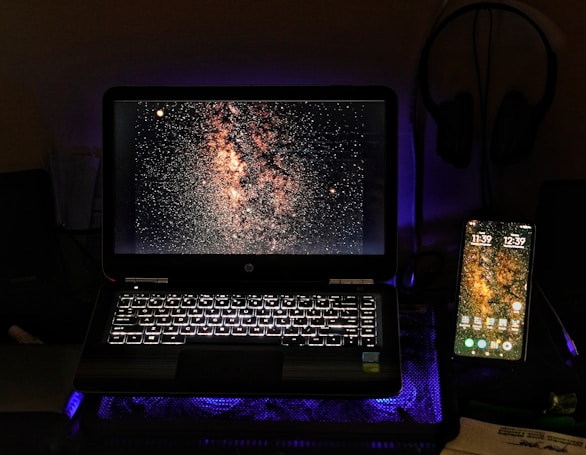 A laptop and a smartphone are displayed side by side on a dark surface. Both screens show an image of a starry sky, possibly the Milky Way. The laptop's keyboard is illuminated with a white backlight, adding a soft glow to the scene. A pair of headphones is visible in the background, hanging on the wall.