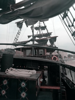 A detailed view of a sailing ship's deck featuring multiple sails tied to masts and rigging. Coiled ropes are neatly placed on a wooden surface, and there is a visible lifebuoy attached to the railing. The ship's elements are clad in dark materials, suggesting an aged or rustic appearance.