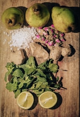 A flat lay of fresh ingredients on a wooden surface. The top row features three whole pears. Below them are cubes of sugar and a cluster of dried rosebuds. In the middle, there are pieces of fresh ginger root. The lower part of the composition shows a bunch of fresh mint leaves, and two halves of a lemon are positioned at the bottom.