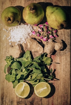 A flat lay of fresh ingredients on a wooden surface. The top row features three whole pears. Below them are cubes of sugar and a cluster of dried rosebuds. In the middle, there are pieces of fresh ginger root. The lower part of the composition shows a bunch of fresh mint leaves, and two halves of a lemon are positioned at the bottom.