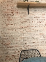A textured whitewashed brick wall with a wooden shelf attached, holding small potted plants. In front, a wire mesh chair partially visible near a table with a mosaic-patterned surface.