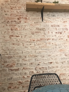 A textured whitewashed brick wall with a wooden shelf attached, holding small potted plants. In front, a wire mesh chair partially visible near a table with a mosaic-patterned surface.