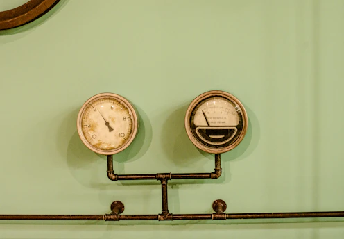 A pair of vintage-style gauges attached to a metal pipe system against a pale green background. The left gauge appears to be a pressure meter, while the right one displays increments and some text, likely indicating measurements.