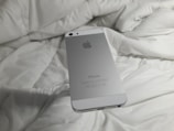 A silver smartphone with an Apple logo rests on a white, rumpled fabric surface. The back of the phone is visible with clear branding.