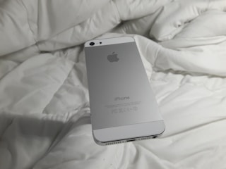 A silver smartphone with an Apple logo rests on a white, rumpled fabric surface. The back of the phone is visible with clear branding.