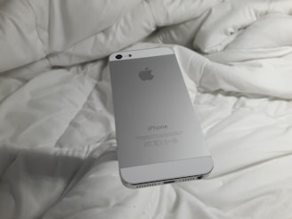 A silver smartphone with an Apple logo rests on a white, rumpled fabric surface. The back of the phone is visible with clear branding.