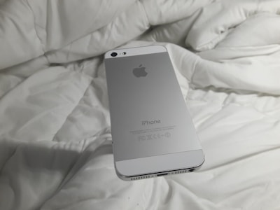 A silver smartphone with an Apple logo rests on a white, rumpled fabric surface. The back of the phone is visible with clear branding.
