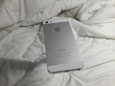 A silver smartphone with an Apple logo rests on a white, rumpled fabric surface. The back of the phone is visible with clear branding.