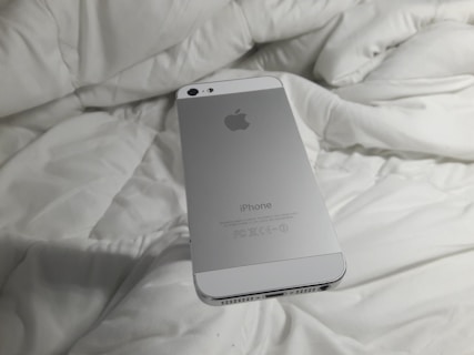A silver smartphone with an Apple logo rests on a white, rumpled fabric surface. The back of the phone is visible with clear branding.