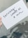 A hand is holding a white card with black handwritten text that reads 'Surviving counts as a win'. The background appears slightly blurred with plant shadows visible.