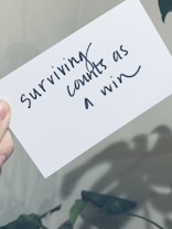 A hand is holding a white card with black handwritten text that reads 'Surviving counts as a win'. The background appears slightly blurred with plant shadows visible.