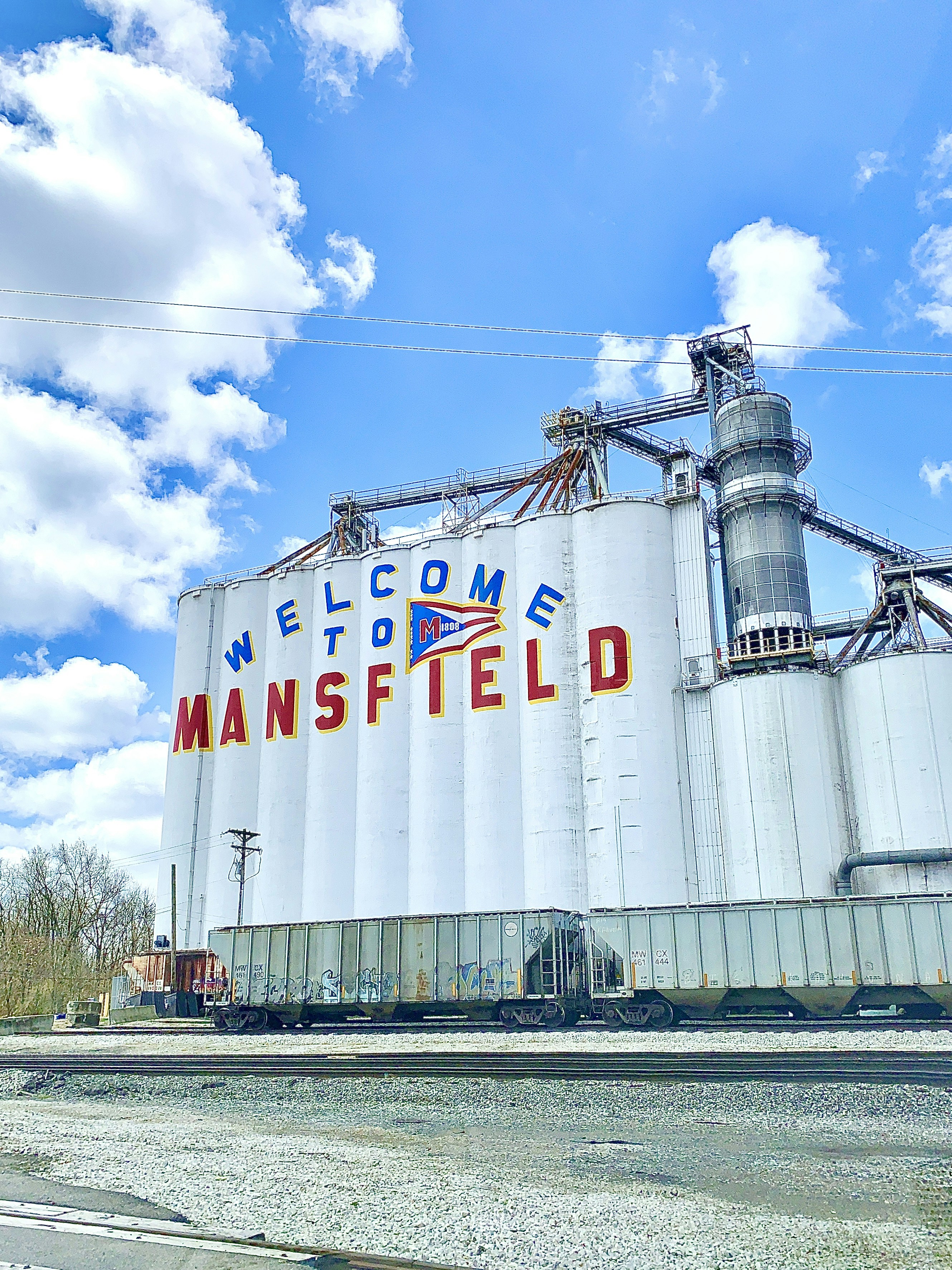Photograph of large grain silos painted with a “Welcome to Mansfield” sign in Mansfield, Ohio, taken on location by the photographer. The image shows industrial infrastructure alongside railroad cars under an open sky, presenting a bold statement of place and local identity. The composition emphasizes scale, signage, and everyday industry, making the image suitable for editorial, regional storytelling, infrastructure, and Americana-themed contexts. The sign’s simplicity allows it to be read broadly as a symbol of arrival and place.