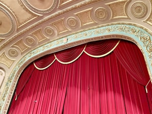 An ornate theater stage with a rich red curtain drawn, set against a detailed and decorative ceiling featuring gold and cream intricate moldings and carvings.