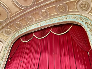 An ornate theater stage with a rich red curtain drawn, set against a detailed and decorative ceiling featuring gold and cream intricate moldings and carvings.