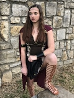 A person dressed in a medieval or fantasy-style costume stands against a stone wall. The outfit includes a dark top with gold trim, a maroon cape, and gladiator-style sandals. The person's long brown hair is adorned with a simple headband, and they wear dark red lipstick.