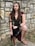 A person dressed in a medieval or fantasy-style costume stands against a stone wall. The outfit includes a dark top with gold trim, a maroon cape, and gladiator-style sandals. The person's long brown hair is adorned with a simple headband, and they wear dark red lipstick.