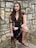 A person dressed in a medieval or fantasy-style costume stands against a stone wall. The outfit includes a dark top with gold trim, a maroon cape, and gladiator-style sandals. The person's long brown hair is adorned with a simple headband, and they wear dark red lipstick.