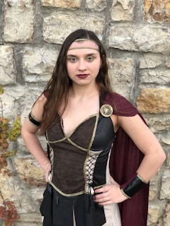 A person dressed in a medieval-inspired costume stands against a stone wall. The costume includes a black and gold bodice with a dark red cape, a gold headband, and arm cuffs. The wall is made of large, roughly-cut stones with some areas of vegetation and vines growing on it.