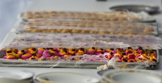 A table is lined with trays containing colorful floral arrangements covered in plastic wrap. The foreground showcases vibrant pink and orange flowers, while the background has a more subdued tone with other objects.