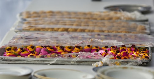 A table is lined with trays containing colorful floral arrangements covered in plastic wrap. The foreground showcases vibrant pink and orange flowers, while the background has a more subdued tone with other objects.