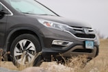 A dark gray Honda CR-V is parked on a grassy and rocky terrain. The car's front license plate reads 'SHIVA.' The weather appears overcast, with a cloudy sky in the background.