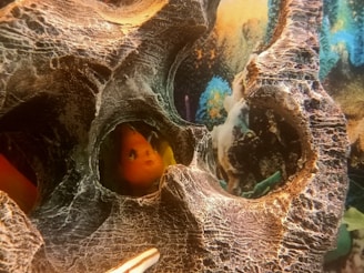 An artificial coral or rock structure with detailed textures, featuring a bright orange goldfish peeking out from a crevice. The vibrant backdrop consists of colorful aquatic plants and abstract underwater scenery, adding depth and intrigue to the composition.